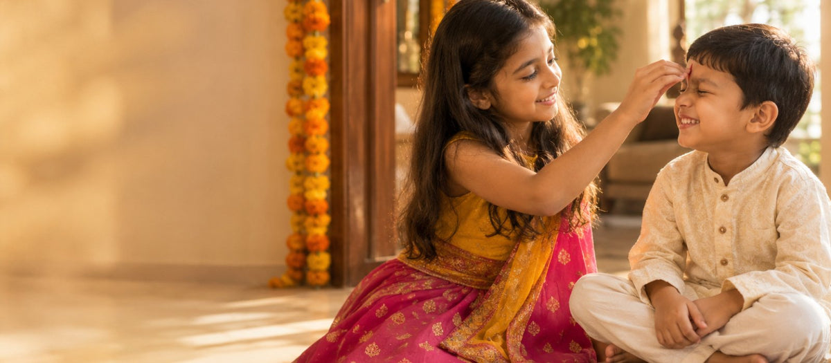 A young Indian girl in a pink lehenga applies a tilak to her little brother's forehead on Bhai Dooj, with an aarti thali, diya and marigolds
