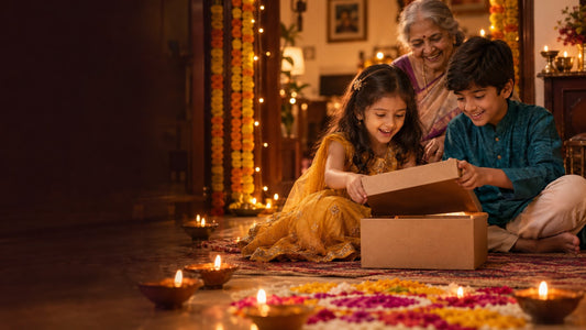 Brother and sister in festive clothes opening a gift box together on Diwali evening while their grandmother smiles, with diyas, rangoli and a marigold toran